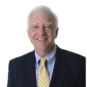 A smiling individual wearing a dark blazer, light blue shirt, and polka dot tie stands against a white background.