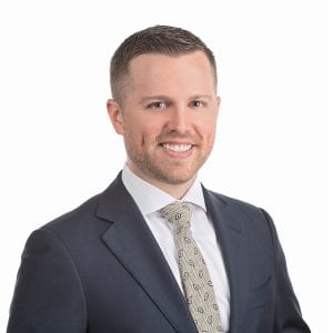 A smiling man in a dark suit and patterned tie stands against a white background.