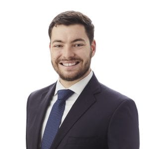 A man with a beard is smiling and wearing a suit and tie against a white background.