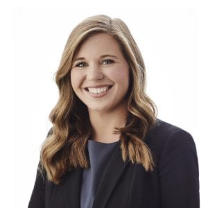 A person with long, wavy hair is smiling, wearing a dark blazer over a gray top, against a white background.