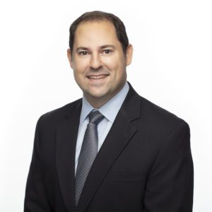 A person wearing a dark suit, light blue shirt, and tie smiles against a white background.