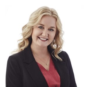 A smiling woman with wavy blonde hair is wearing a dark blazer over a red blouse against a plain white background.