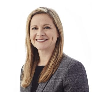 A smiling individual with long blonde hair is wearing a grey blazer and a black top against a white background.