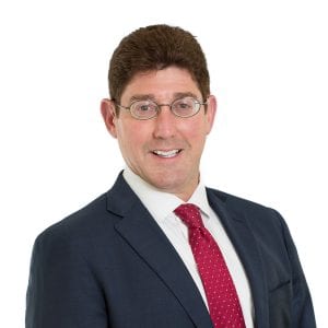 A man wearing a suit, red tie, and glasses is smiling against a white background.