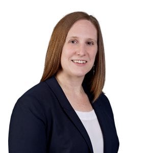 A person with shoulder-length brown hair is wearing a black blazer over a white top and smiling at the camera.