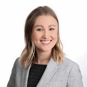 A person with shoulder-length blonde hair is smiling and wearing a gray blazer over a black top.