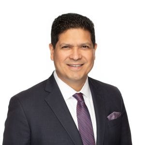 A person in a suit and tie is smiling against a plain white background.