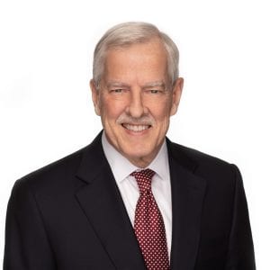 A man with gray hair is smiling and wearing a dark suit with a white shirt and a red tie with white polka dots.