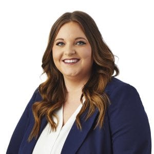 A woman with long, wavy hair smiles while wearing a navy blazer over a white shirt against a plain background.