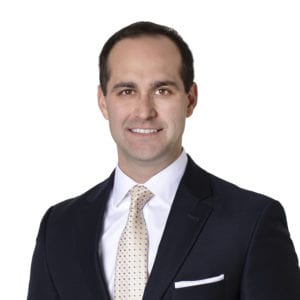 A man in a dark suit with a white dress shirt and a polka dot tie smiles against a white background.