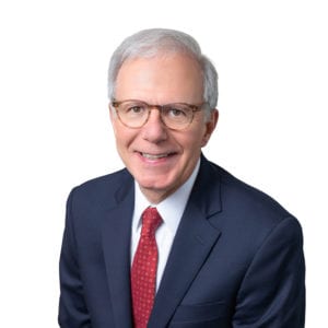 An elderly individual with white hair is wearing glasses, a dark suit, a white shirt, and a red tie, smiling against a white background.