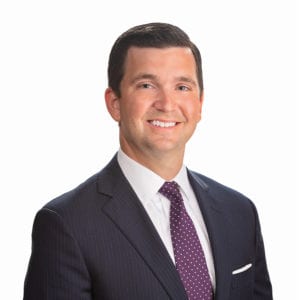 A man in a dark suit with a white shirt and a purple patterned tie is smiling at the camera.