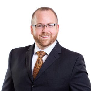 A man with a beard and glasses is wearing a dark suit and an orange patterned tie, smiling against a white background.