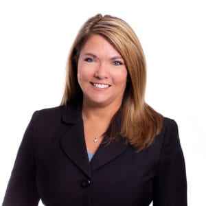 A woman with long, light brown hair is wearing a dark blazer and smiling against a white background.