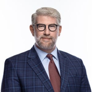 A man with glasses, gray hair, and a beard is wearing a plaid suit jacket, light blue shirt, and burgundy tie, posing against a plain white background.