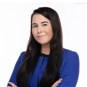 A person with long, dark hair is wearing a blue jacket and smiling with their arms crossed against a white background.