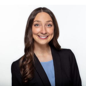 A smiling person with long brown hair wearing a black blazer and a light blue top stands against a plain white background.