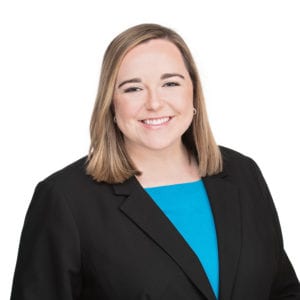 A person with shoulder-length hair wearing a black blazer and a blue top is smiling against a white background.
