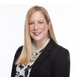A woman with long blonde hair, wearing a black blazer over a patterned blouse and a pearl necklace, smiles at the camera against a plain white background.