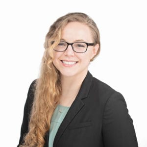A smiling person with long, wavy hair and glasses is wearing a black blazer and a green top against a white background.