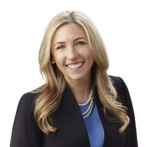 A smiling person with long blonde hair is dressed in business attire, including a black blazer, blue top, and a pearl necklace, against a white background.