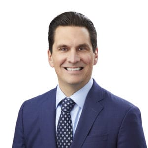 A person wearing a blue suit and patterned tie is smiling against a plain background.