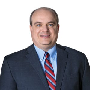 A person is wearing a dark suit with a light blue shirt and red and blue striped tie, smiling at the camera against a white background.