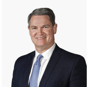 A person wearing a dark suit, white shirt, and blue tie is smiling against a plain white background.