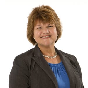 A woman with short brown hair, wearing a gray blazer over a blue top and a necklace, is smiling against a white background.