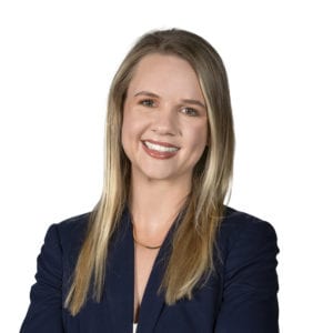 A person with long straight blond hair wearing a navy blue blazer and a white top smiles while standing against a plain white background.