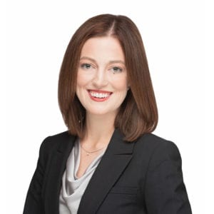 A person with straight, shoulder-length brown hair is smiling and wearing a professional black blazer over a grey blouse.