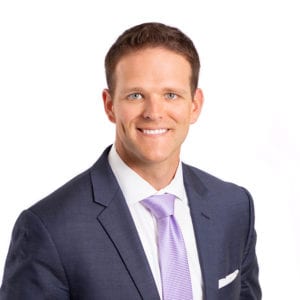 A person is wearing a suit with a white shirt and a light purple tie, smiling against a white background.