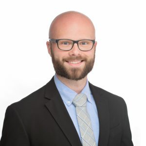 A bearded man with glasses is wearing a suit and tie and smiling against a plain white background.