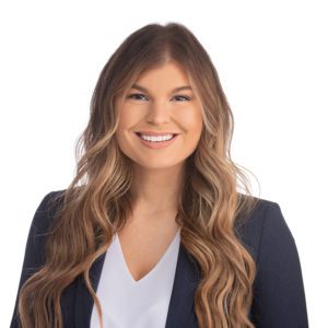 A person with long, wavy hair, wearing a white blouse and a dark blazer, smiles at the camera against a plain background.