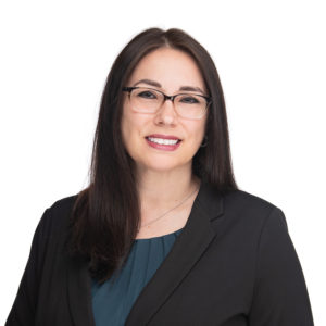 A person with long dark hair and glasses is dressed in a black blazer and teal top, smiling against a white background.