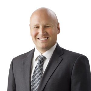 A bald man in a suit and tie is smiling against a plain white background.