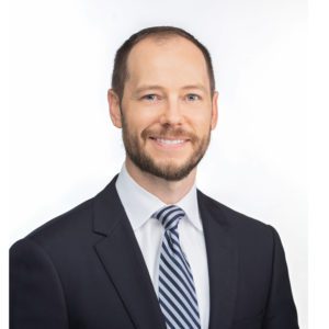A man wearing a suit and striped tie is smiling against a plain white background.