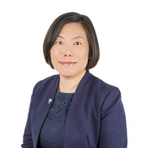 A person wearing a dark blazer and a blue top is looking at the camera against a white background.