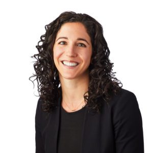 A person with curly dark hair is smiling while wearing a black blazer and a dark top in front of a white background.