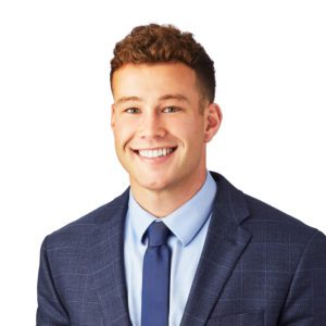 A smiling person wearing a blue suit, light blue dress shirt, and navy tie against a white background.