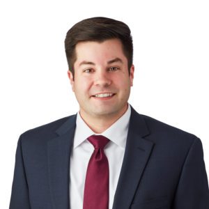 A man in a dark suit, white shirt, and red tie is smiling.