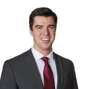 A well-dressed man in a dark suit and red tie is smiling against a plain white background.