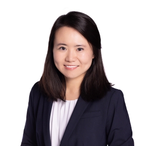 A woman with shoulder-length dark hair is wearing a dark blazer and a white top, smiling against a plain white background.