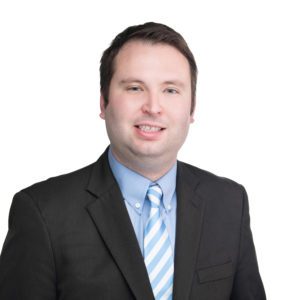 A man in a black suit and blue striped tie stands smiling against a white background.
