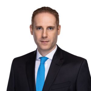 A person wearing a black suit with a blue tie, posed against a white background.