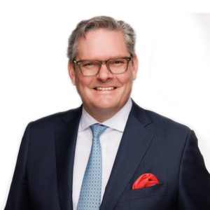 A person wearing a navy suit, light blue tie, and glasses, with a red pocket square, smiles against a white background.