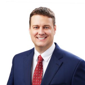 A man is dressed in a blue suit, white shirt, and red patterned tie, smiling against a plain white background.