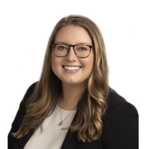 A smiling woman with long, wavy hair wearing glasses and professional attire.