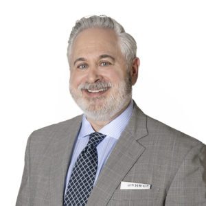 A man with gray hair and beard is dressed in a suit and tie, smiling against a white background.