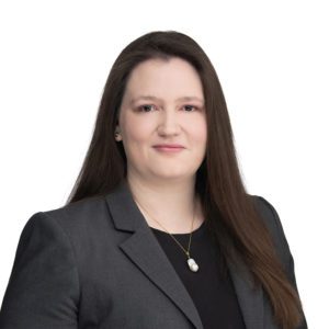 A person with long hair is wearing a black top and grey blazer, accessorized with a small necklace featuring a pearl.